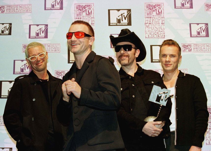 Czterech mężczyzn w okularach przeciwsłonecznych, pozujący z nagrodą MTV na tle plakatu.