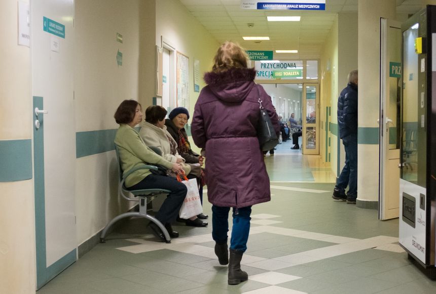 Korytarz w przychodni z pacjentami siedzącymi na ławkach, idąca kobieta w płaszczu.
