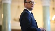 Mężczyzna w garniturze, odwrócony w stronę kamery, na tle eleganckiego wnętrza.