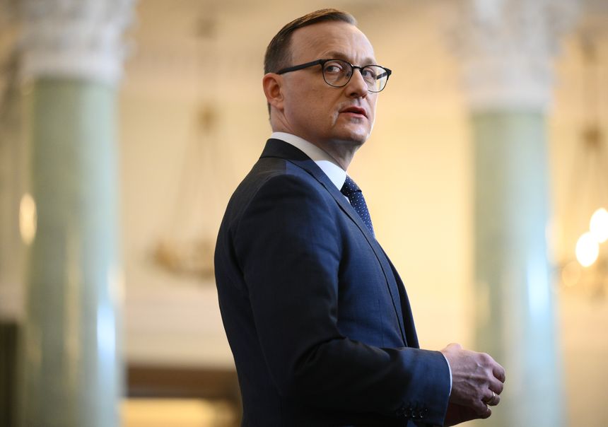 Mężczyzna w garniturze, odwrócony w stronę kamery, na tle eleganckiego wnętrza.