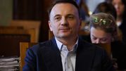 Mężczyzna w garniturze siedzi w sali, z uśmiechem na twarzy. W tle widać inne osoby.