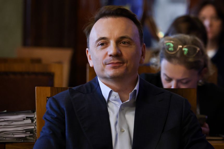 Mężczyzna w garniturze siedzi w sali, z uśmiechem na twarzy. W tle widać inne osoby.