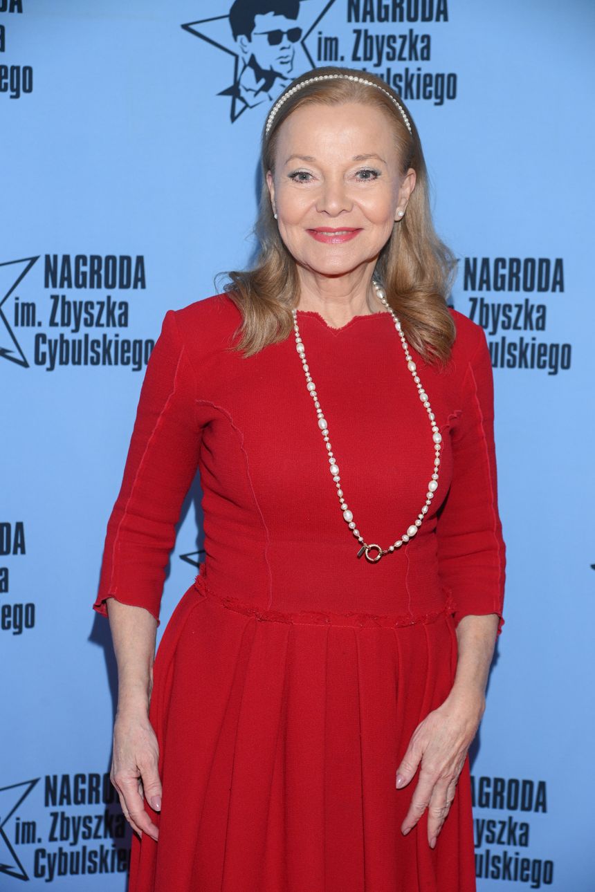Kobieta w czerwonej sukience z perłami na szyi, uśmiechająca się na tle niebieskiego banera.
