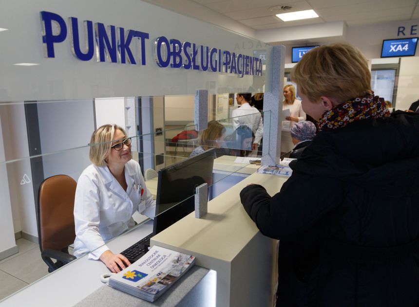 Kobieta w białym fartuchu rozmawia z pacjentką przy punkcie obsługi. W tle inni pracownicy.