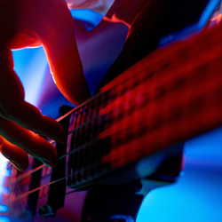 Zdjęcie przedstawia rękę grającą na gitarze basowej w intensywnym świetle kolorowych reflektorów.