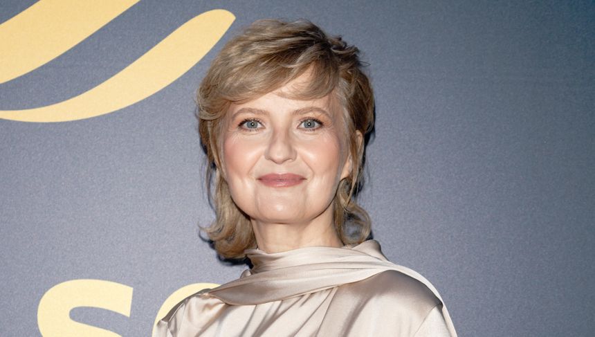 Izabela Kuna o jasnych włosach w bluzce w kolorze beżowym, uśmiecha się na tle ciemnej ściany.