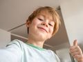 Chłopiec z uśmiechem robi sobie selfie, trzymając kciuk w górze na tle jasnego wnętrza.