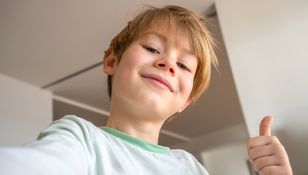 Chłopiec z uśmiechem robi sobie selfie, trzymając kciuk w górze na tle jasnego wnętrza.
