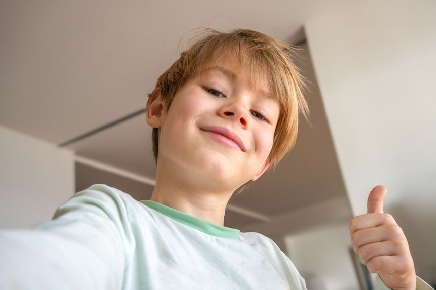 Chłopiec z uśmiechem robi sobie selfie, trzymając kciuk w górze na tle jasnego wnętrza.