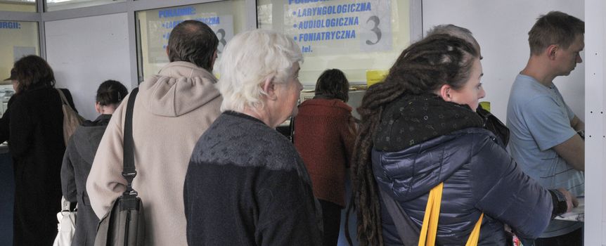 Osoby czekające w kolejce w przychodni, widać numery i gabinety poradni.