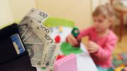 Portfel z banknotami 100 zł, w tle bawiące się dziecko przy stole.