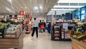 Opis ALT: Wnętrze supermarketu z półkami pełnymi owoców i warzyw, klienci w alejce zakupowej.