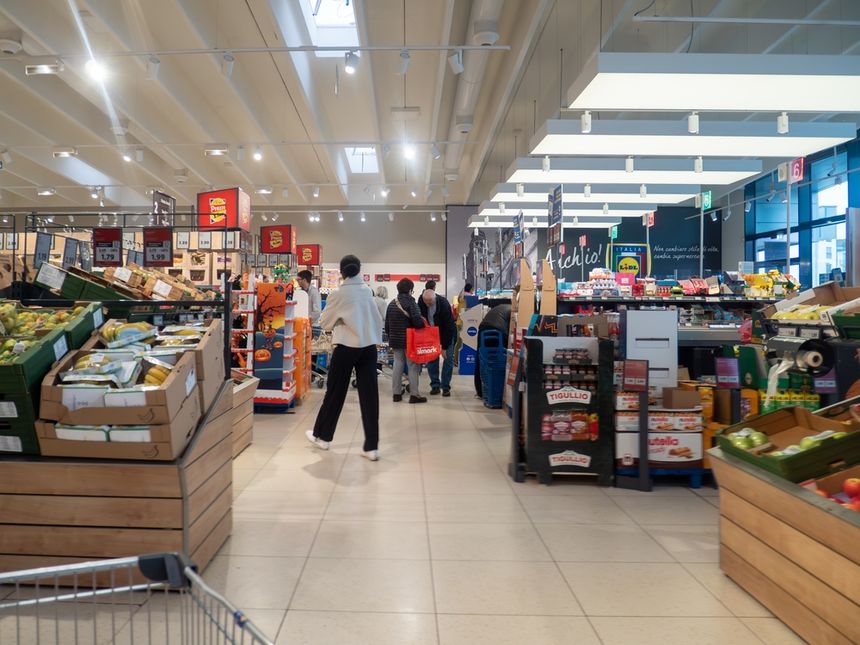 Opis ALT: Wnętrze supermarketu z półkami pełnymi owoców i warzyw, klienci w alejce zakupowej.