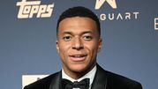 Kylian Mbappe w eleganckim garniturze z czarną muszką na tle ciemnego tła.