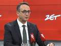 Mężczyzna w garniturze siedzi przy stole, mówi do mikrofonów z logo ZET. Tło czerwone.