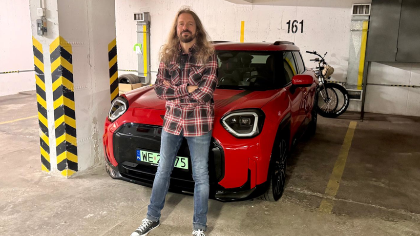 Mężczyzna z długimi włosami w koszuli w kratę stoi obok czerwonego samochodu w parkingu.