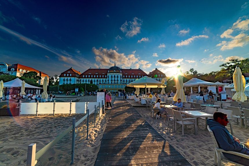 Widok na plażę z drewnianym deptakiem i Grand Hotelem w tle, przy zachodzie słońca.