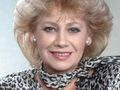Uśmiechnięta kobieta z blond włosami w krzykliwej, wzorzystej bluzce.