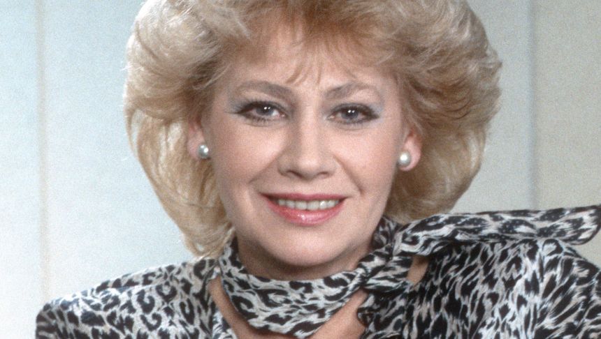 Uśmiechnięta kobieta z blond włosami w krzykliwej, wzorzystej bluzce.