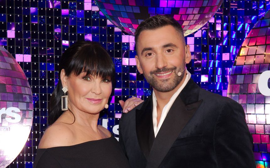 Rafał Maserak i Iwona Pavlović uśmiechają się na tle świecących dekoracji. Oboje są elegancko ubrani.