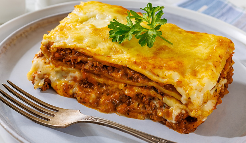 Lasagne na talerzu, warstwy mięsa i sosu, udekorowana gałązką pietruszki. Srebrny widelec obok.