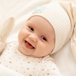 Uśmiechnięte niemowlę w białej czapeczce trzyma pluszowego króliczka. Płaszczyk z gwiazdkami.