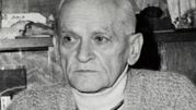 Osoba w starszym wieku w swetrze, z poważnym wyrazem twarzy, siedząca w pomieszczeniu.