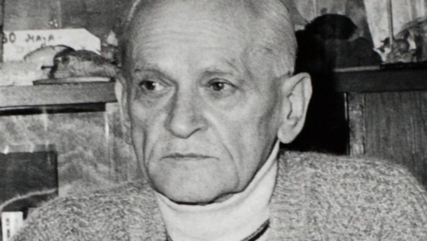 Osoba w starszym wieku w swetrze, z poważnym wyrazem twarzy, siedząca w pomieszczeniu.