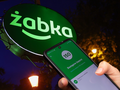 Na zdjęciu zielony szyld Żabki, widać też dłoń trzymającą telefon z aplikacją.
