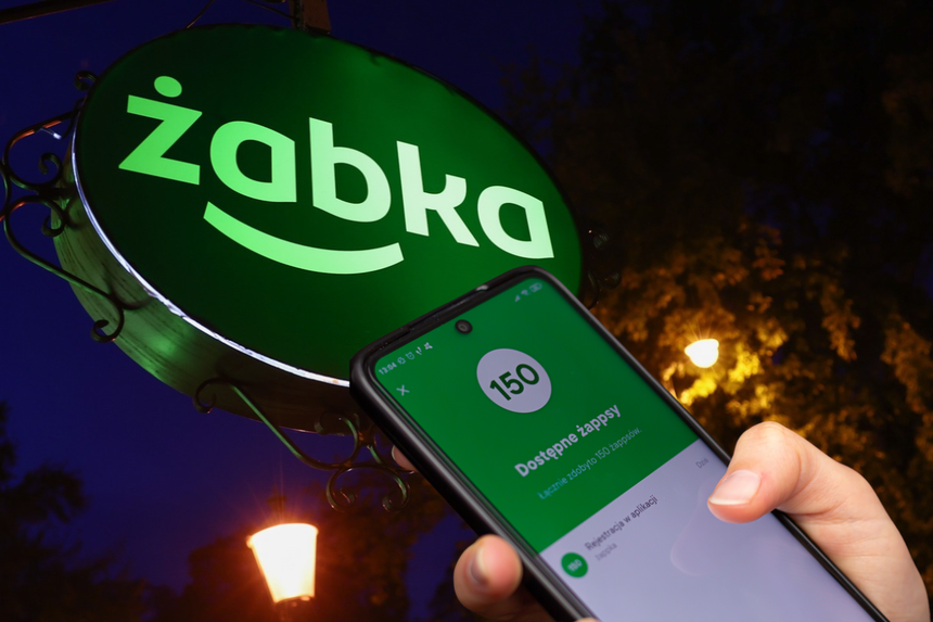 Na zdjęciu zielony szyld Żabki, widać też dłoń trzymającą telefon z aplikacją.