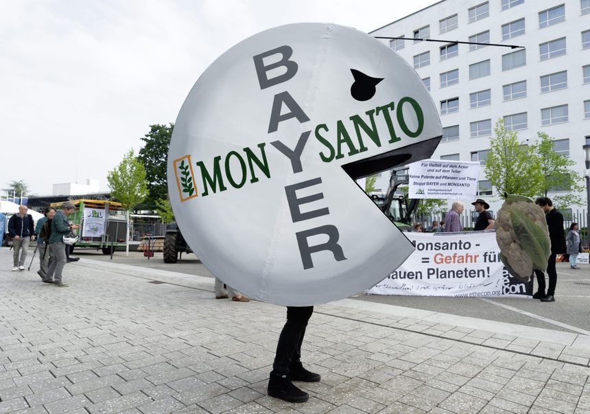 Osoba w kostiumie symbolizującym firmę Bayer/Monsanto, protestująca na ulicy. W tle plakaty.