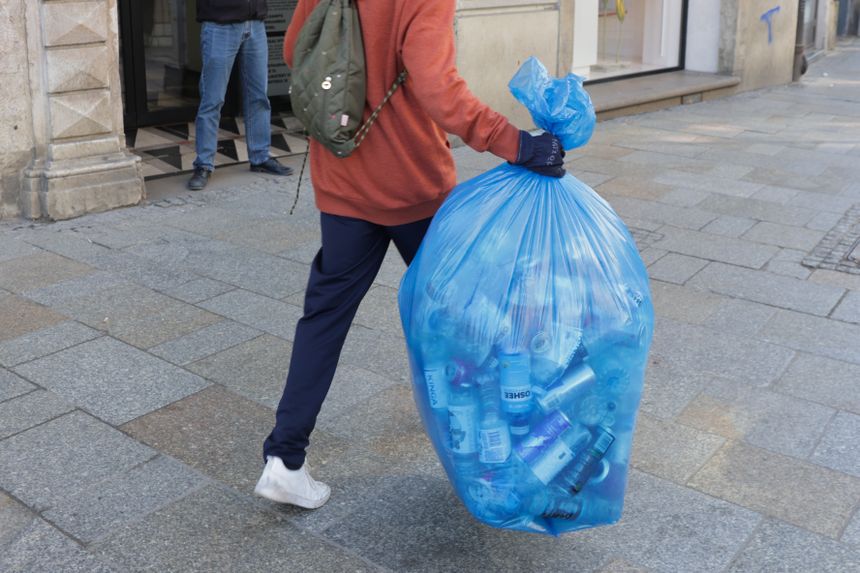 Osoba w czerwonym swetrze niesie dużą niebieską torbę z plastikowymi butelkami w mieście.