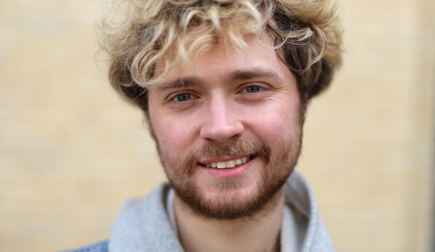 Mężczyzna z kręconymi, blond włosami i brodą, uśmiechający się do kamery na tle jasnej ściany.